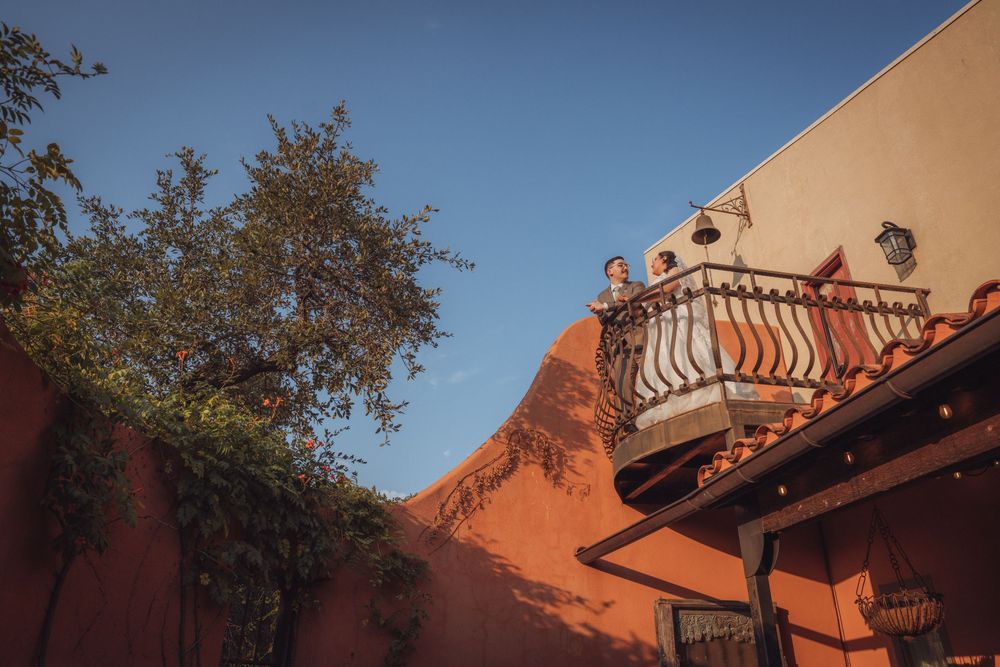 Adobe building with ornate balcony, red rock formation, and lush tree under clear blue sky in Southwest USA.