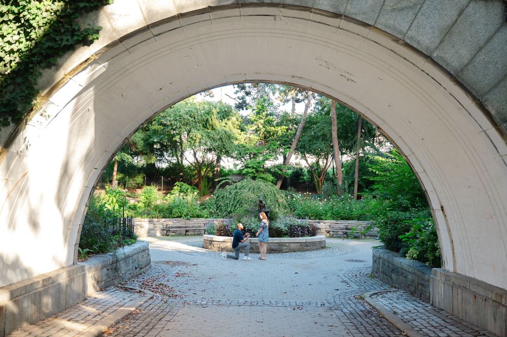 A serene garden viewed through a white stone arch with lush greenery and a person sitting on a bench inside.