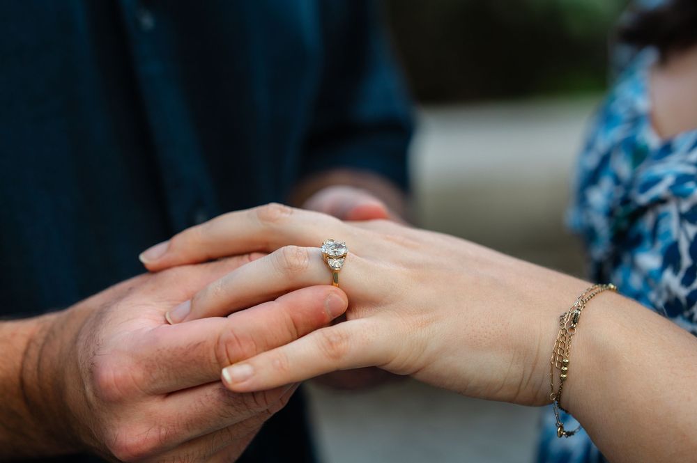 Close-up of an engagement ring on a woman's finger as a man holds her hand outdoors.