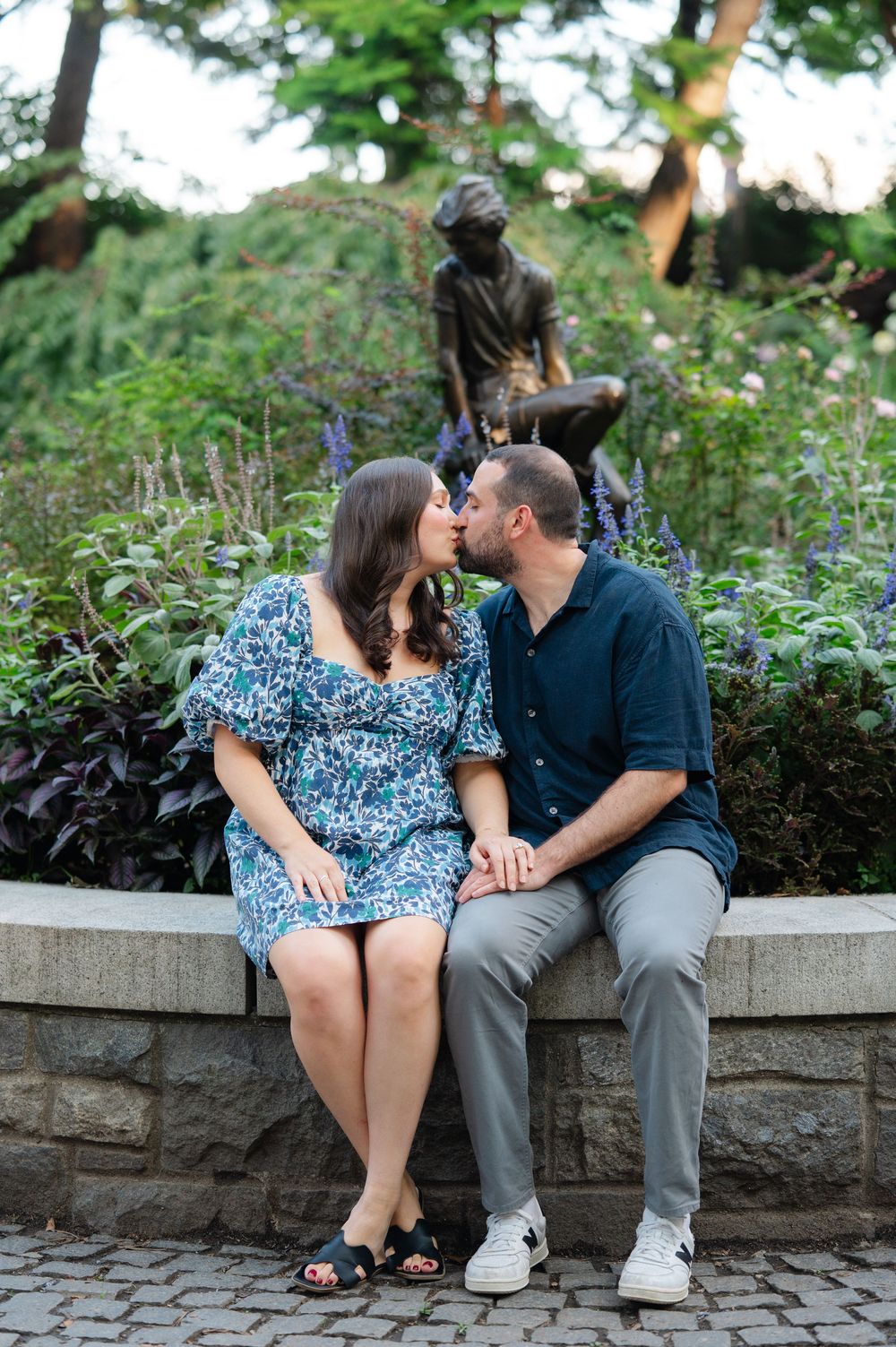 Couple kissing while holding hands on a stone wall surrounded by lush gardens with a bronze statue behind them.