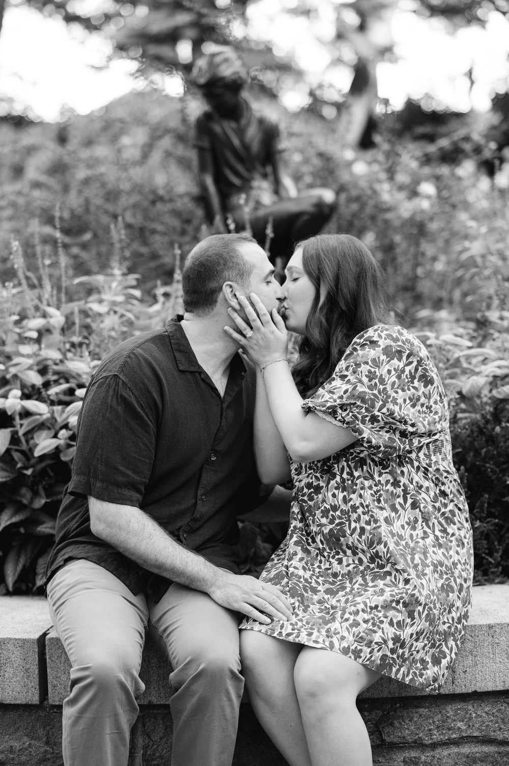 A couple shares a romantic kiss while seated on a stone ledge in a lush garden, with a statue visible behind them.