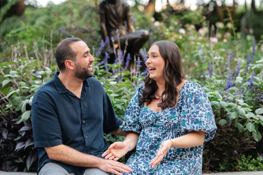 Couple sitting together smiling in a lush garden with purple flowers and bronze sculptures in the background.