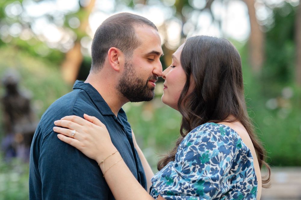 Couple sharing a romantic almost-kiss outdoors, man in blue shirt embracing woman in floral top in a garden setting.