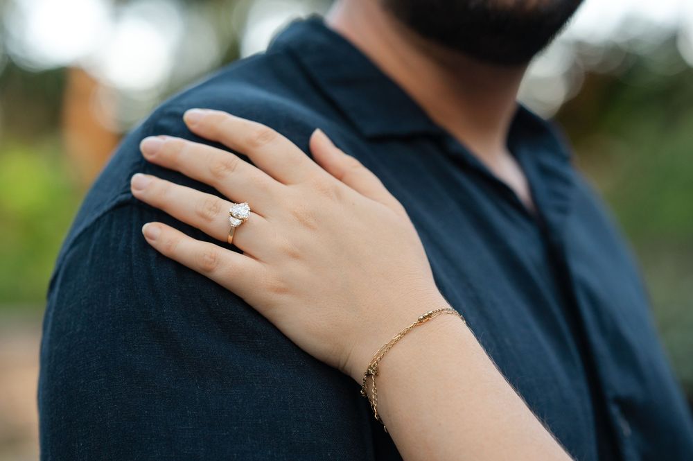 A woman's hand with a diamond engagement ring rests on a man's shoulder, with a gold bracelet visible.