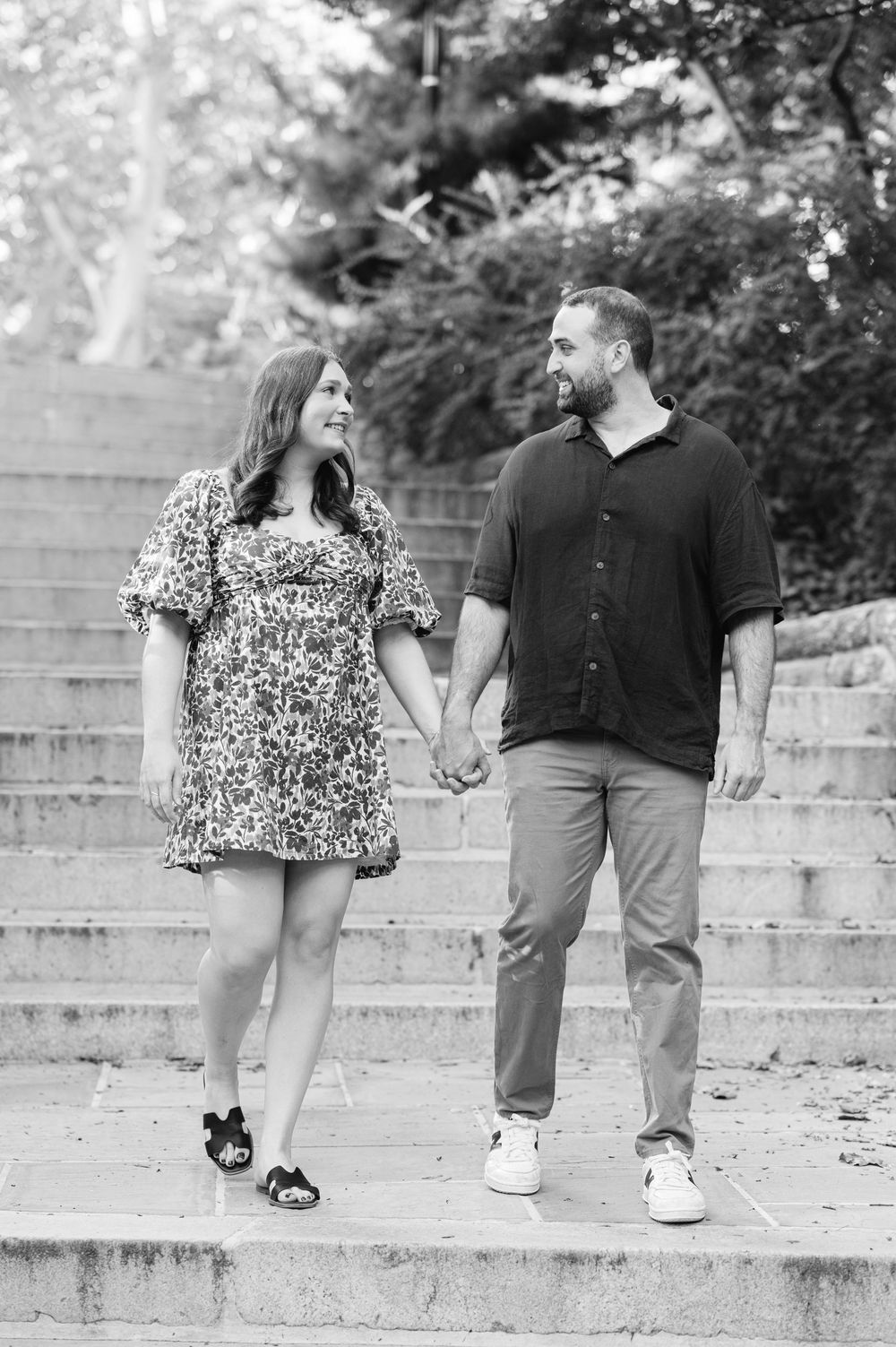 Black and white photo of a couple holding hands and smiling at each other on stone steps outdoors.