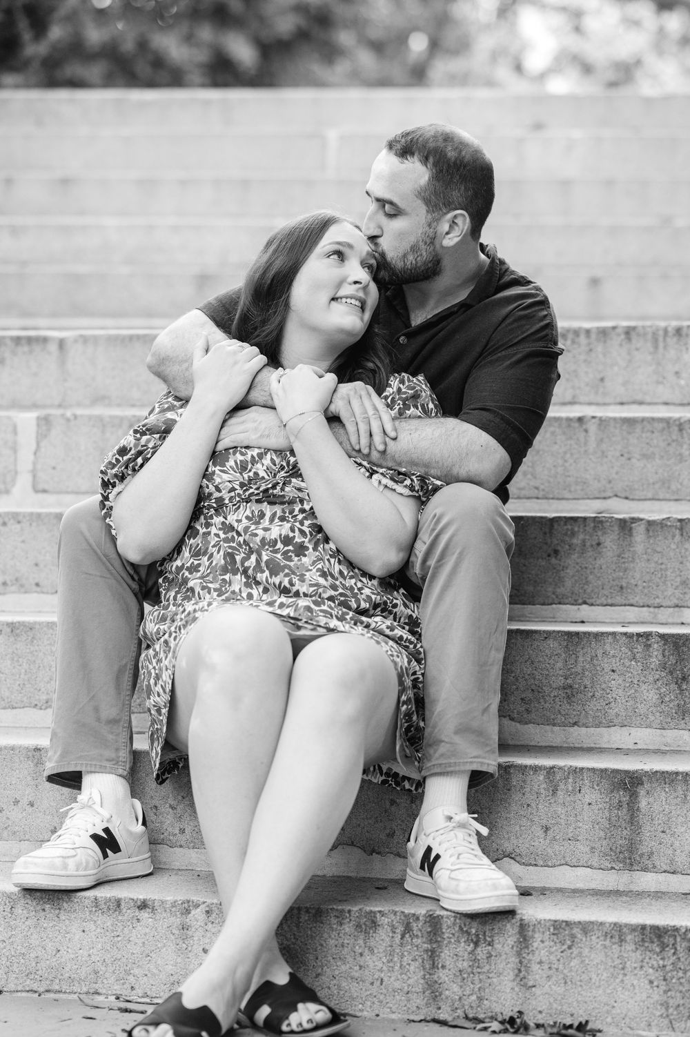 A couple sits on stone steps, the man kissing the woman's cheek as she smiles up at him in a black and white photo.