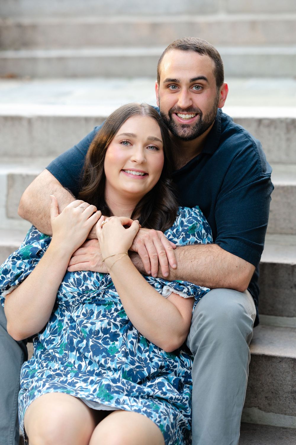 Smiling couple sitting on stone steps, man embracing woman in blue floral dress during engagement photo session.