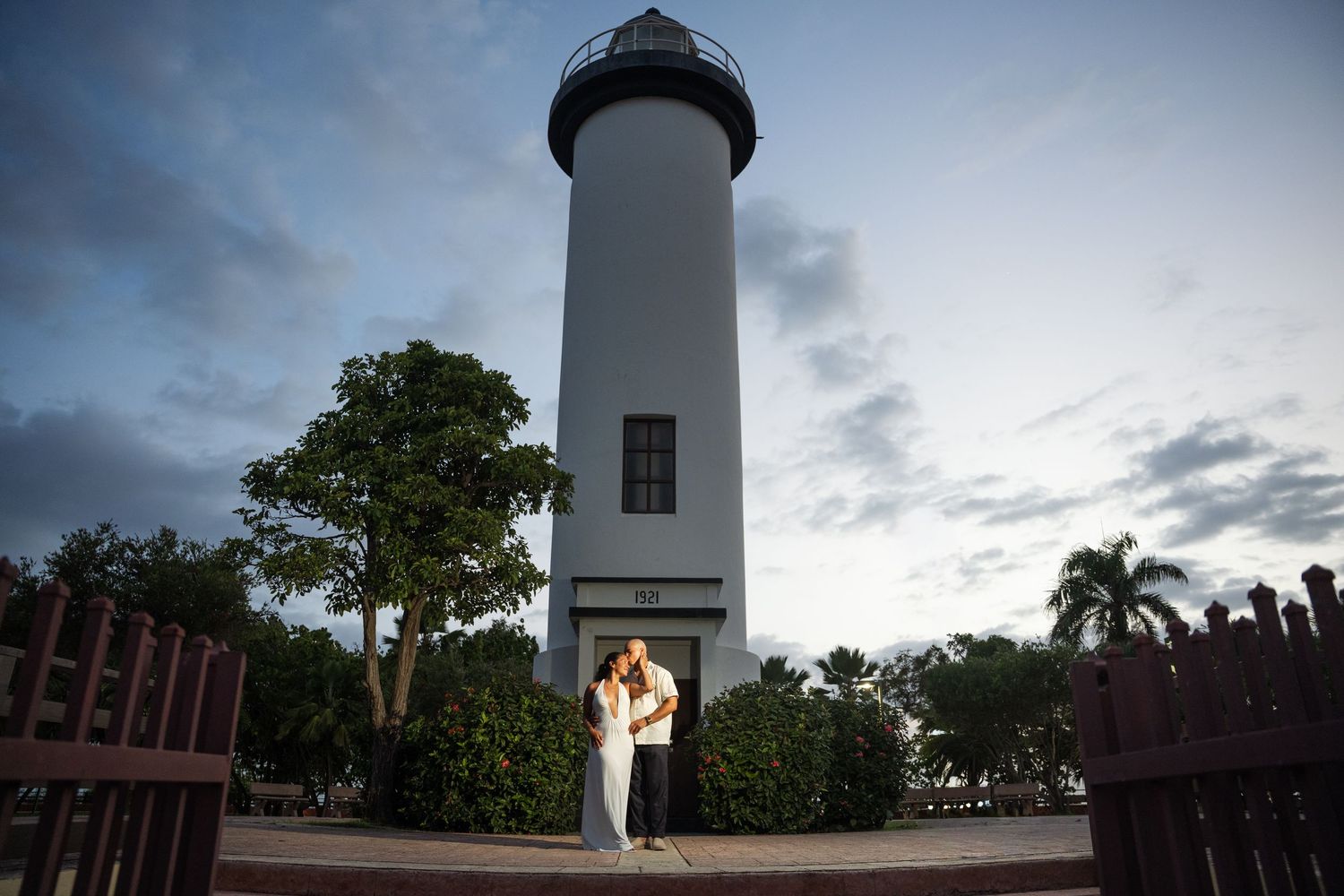 Couple embracing romantically in front of a tall white lighthouse at dusk with palm trees and cloudy sky.