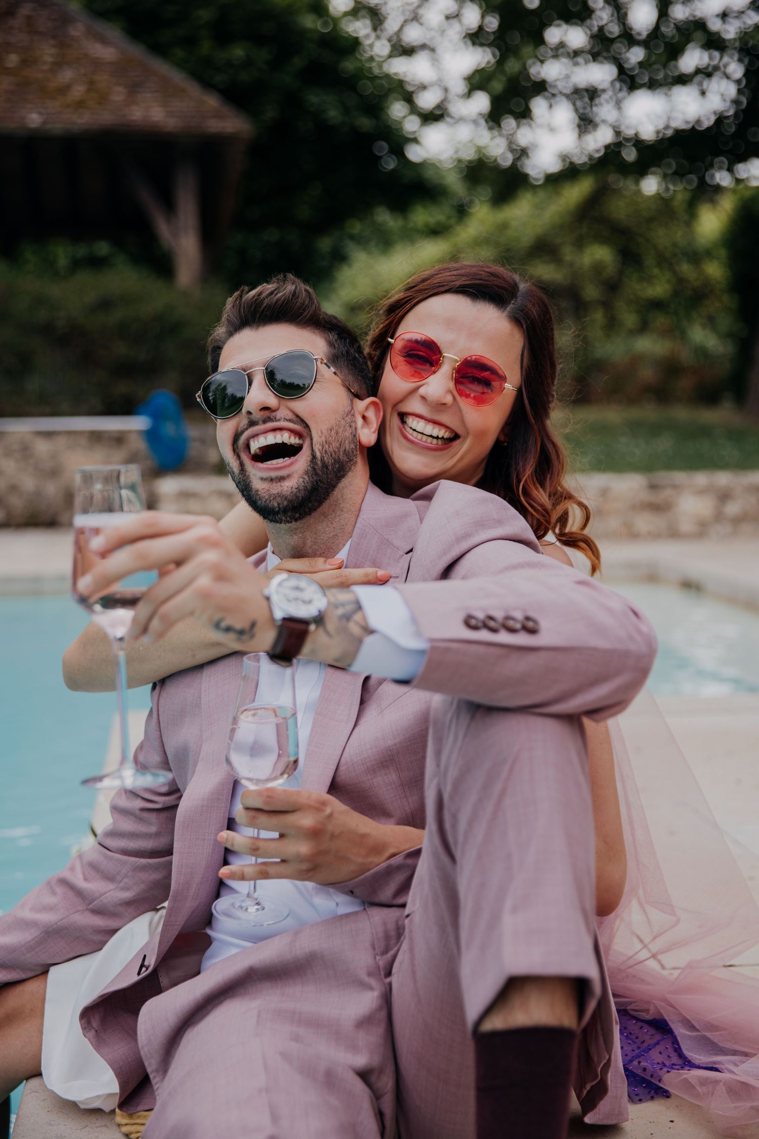 Un couple de jeunes mariés savoure une coupe de champagne en riant, assis au bord de la piscine