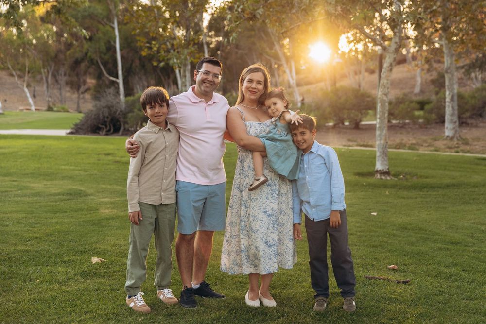 Family of five posing together on green grass in a sunlit park during golden hour.