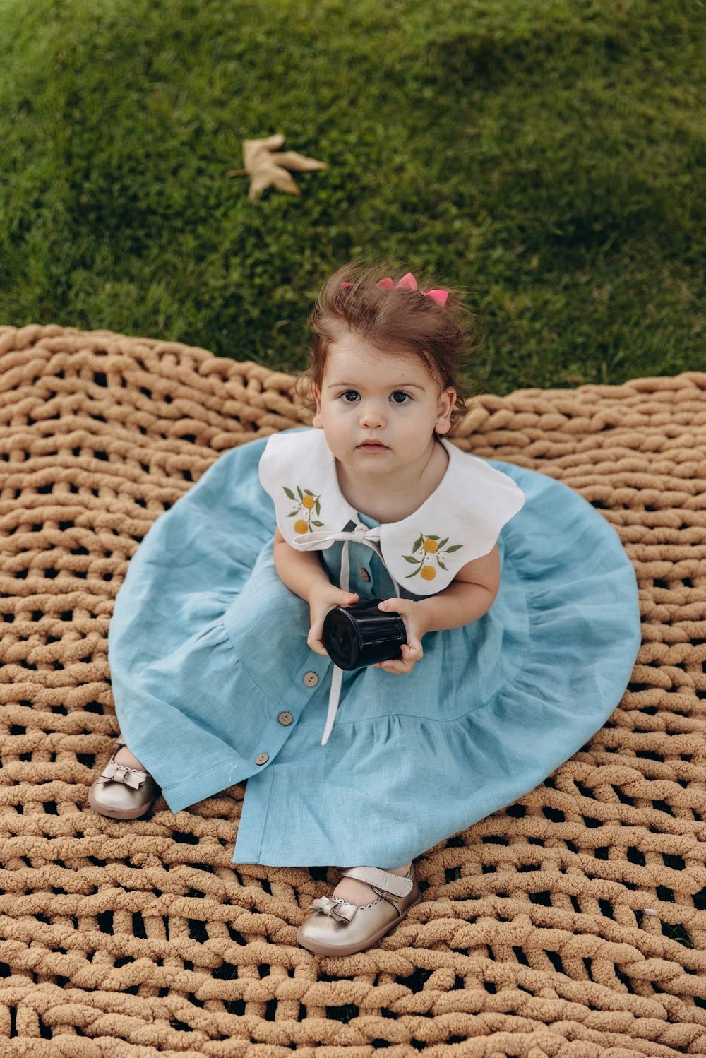 Toddler girl in blue dress with white collar sits on woven mat holding camera, green grass background.