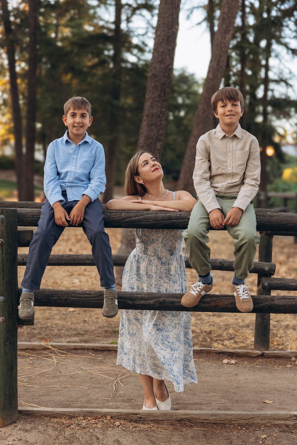 Two boys sit on a rustic wooden fence while a girl in a floral dress stands between them in a park setting.