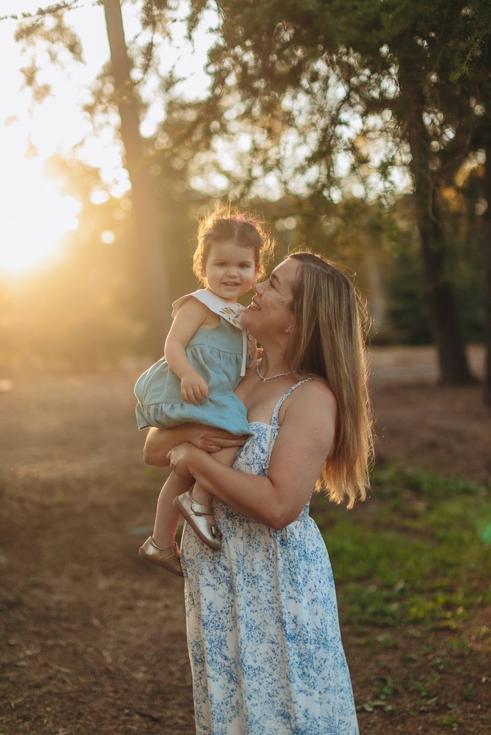A mother in a floral dress holds her toddler in golden sunset light among trees.