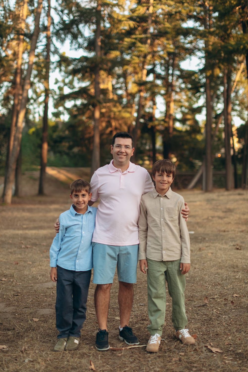 A smiling man with two boys posing together in a sunny pine forest park setting.