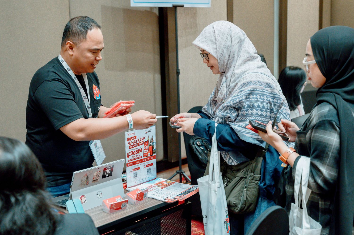 A man in black shirt hands promotional materials to women in hijabs at an event registration booth.