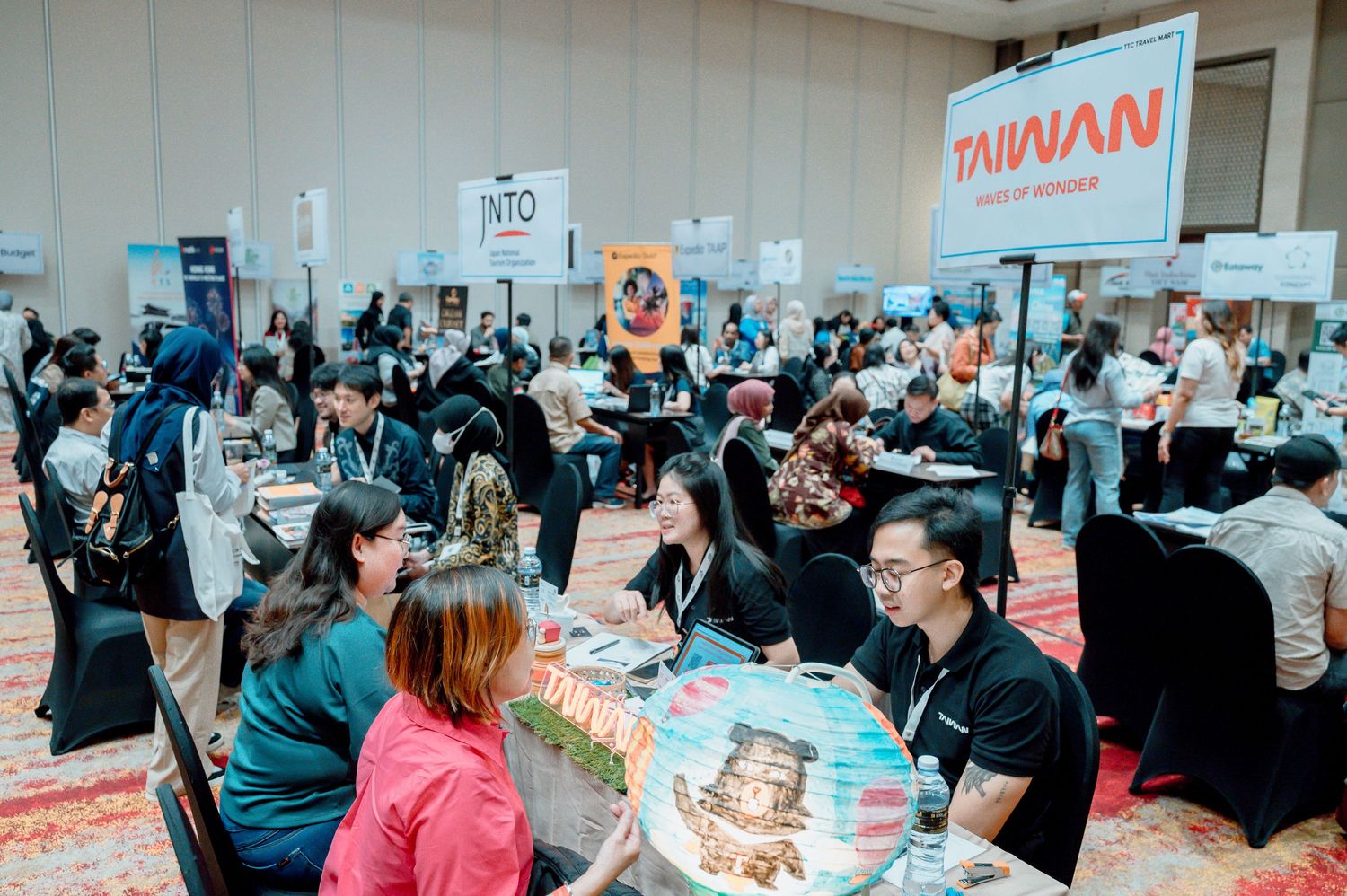 Busy travel expo with Taiwan tourism booth, attendees networking at tables in a large convention hall.