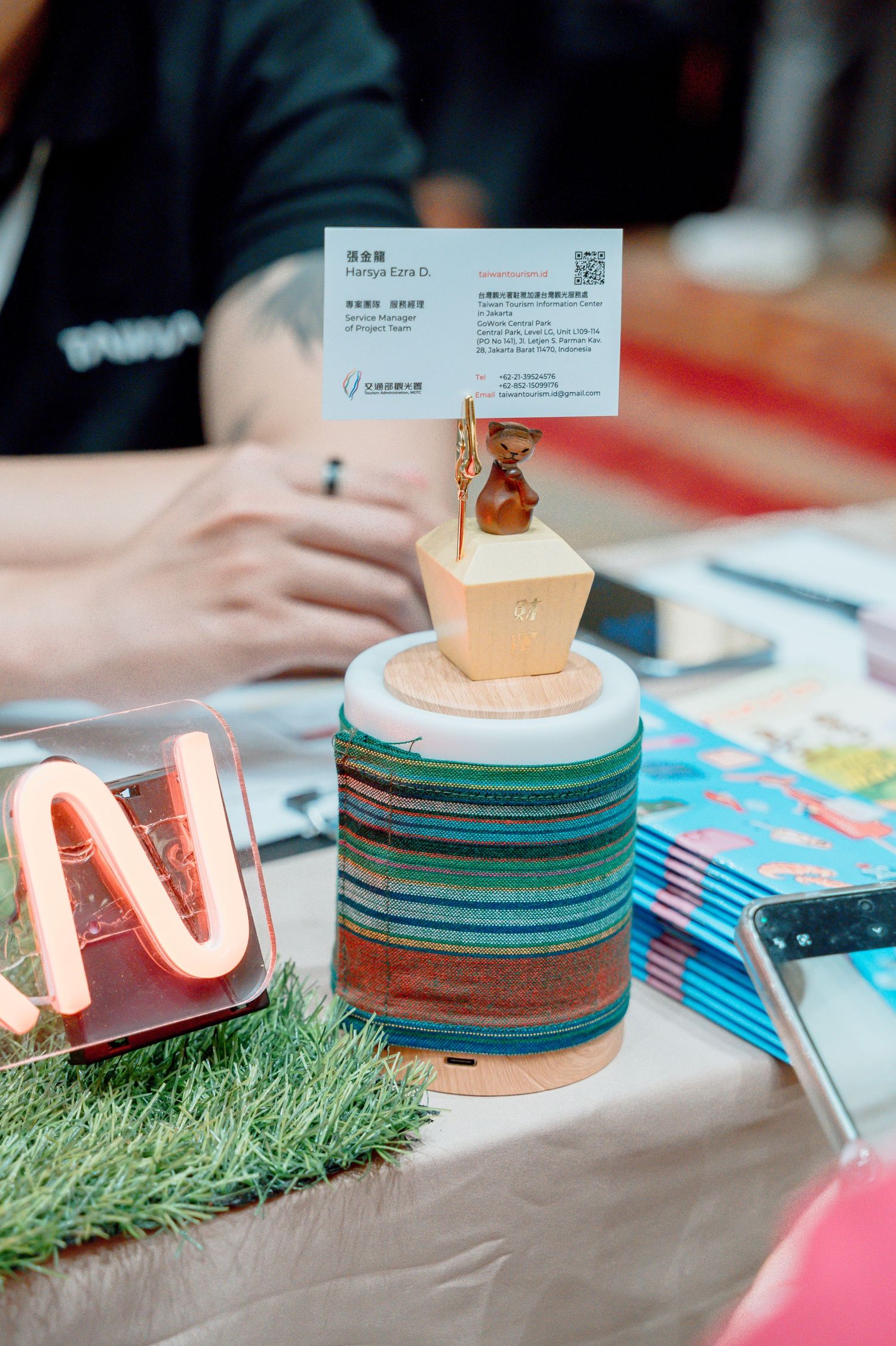 Colorful layered wooden craft object topped with geometric gold figurine displayed on artificial grass at a trade show table.