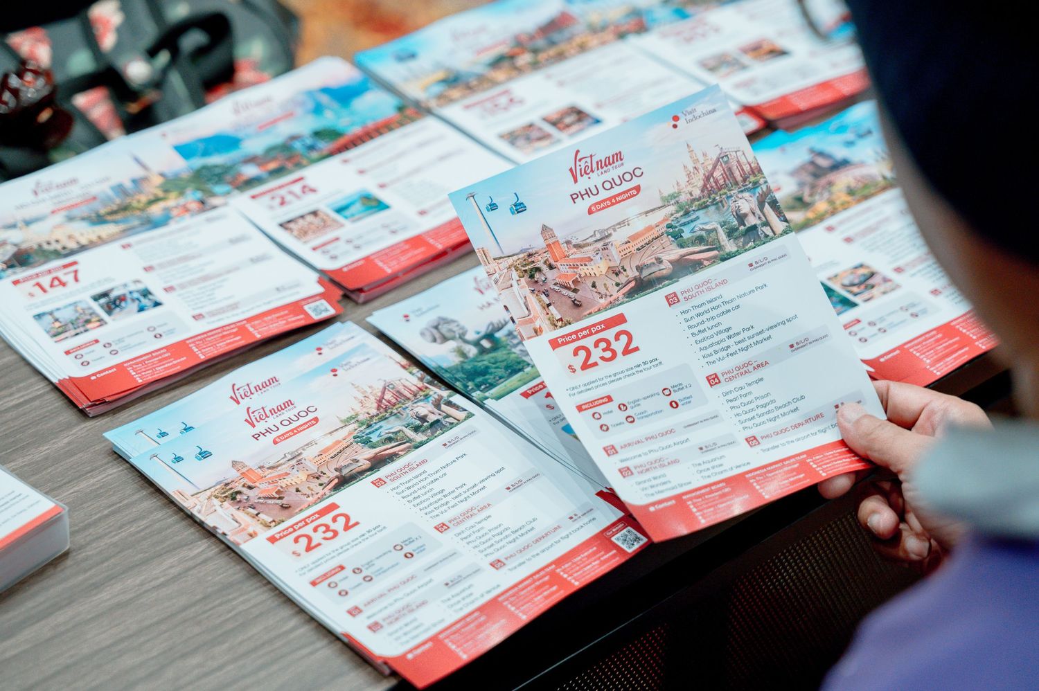 Person browsing colorful Vietnam travel brochures with tour packages priced at 147 and 232 on a wooden table.