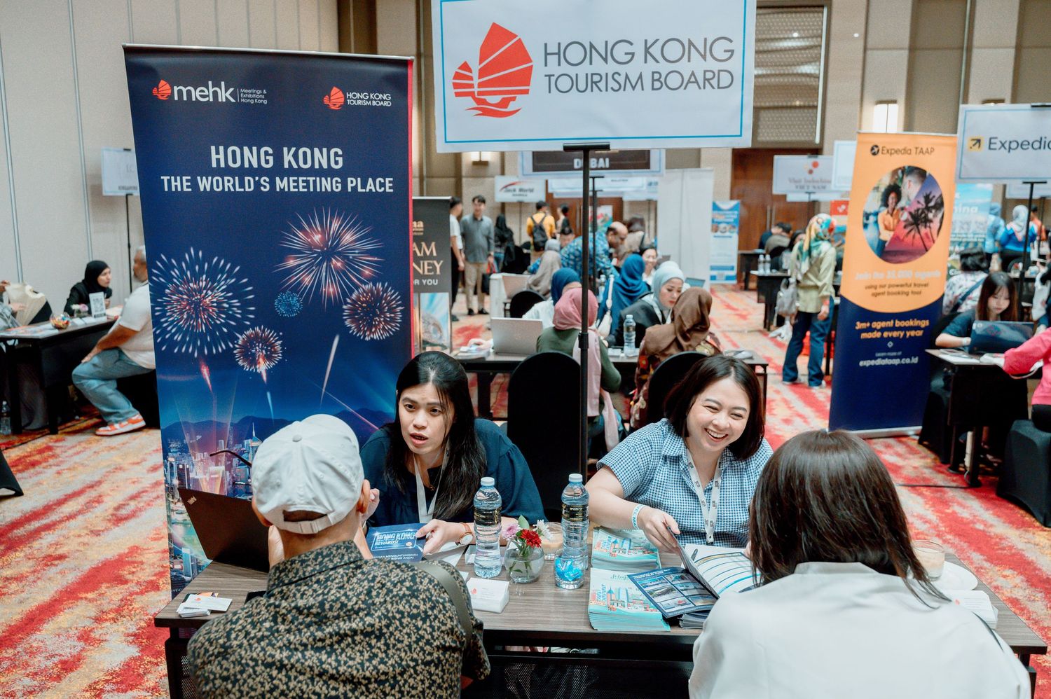 Hong Kong Tourism Board booth at a travel expo with visitors meeting representatives under a colorful banner.