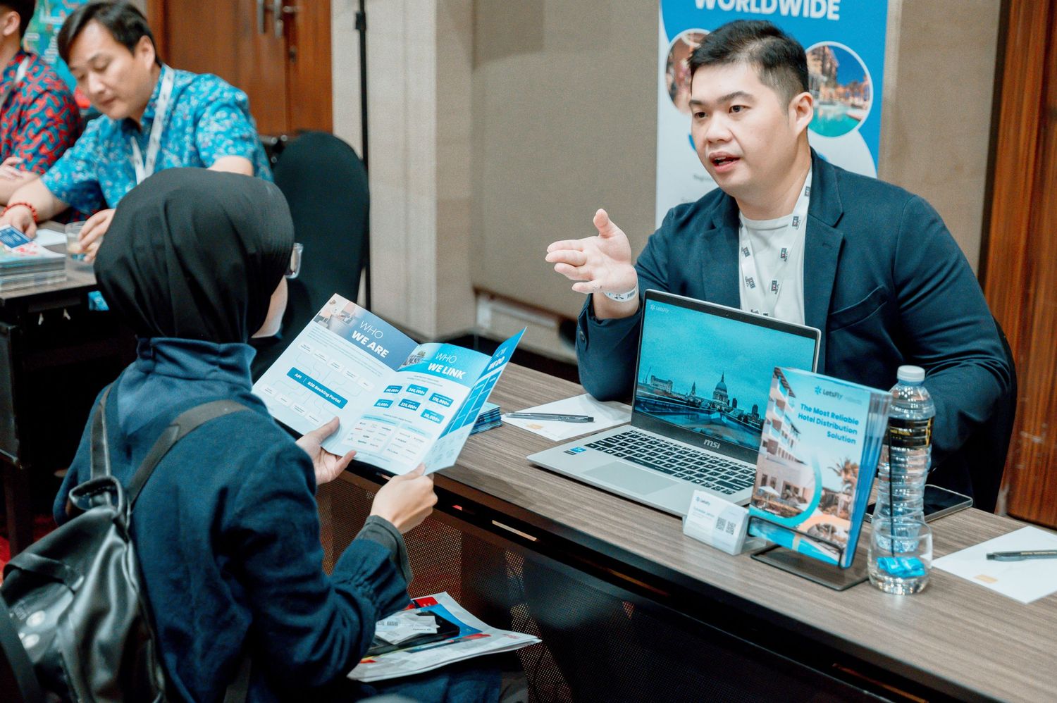 A businessman in a blazer discusses travel brochures with a client at a conference table with a laptop nearby.