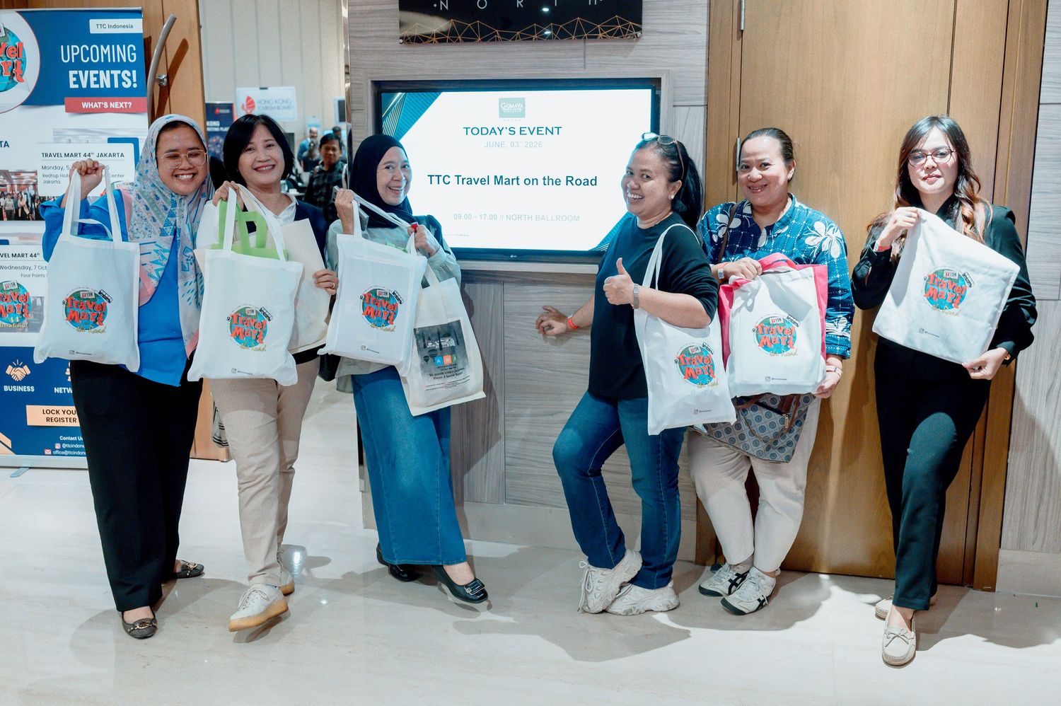 Six women smiling and holding tote bags at a TTC Travel Meet on the Road event in a modern venue.