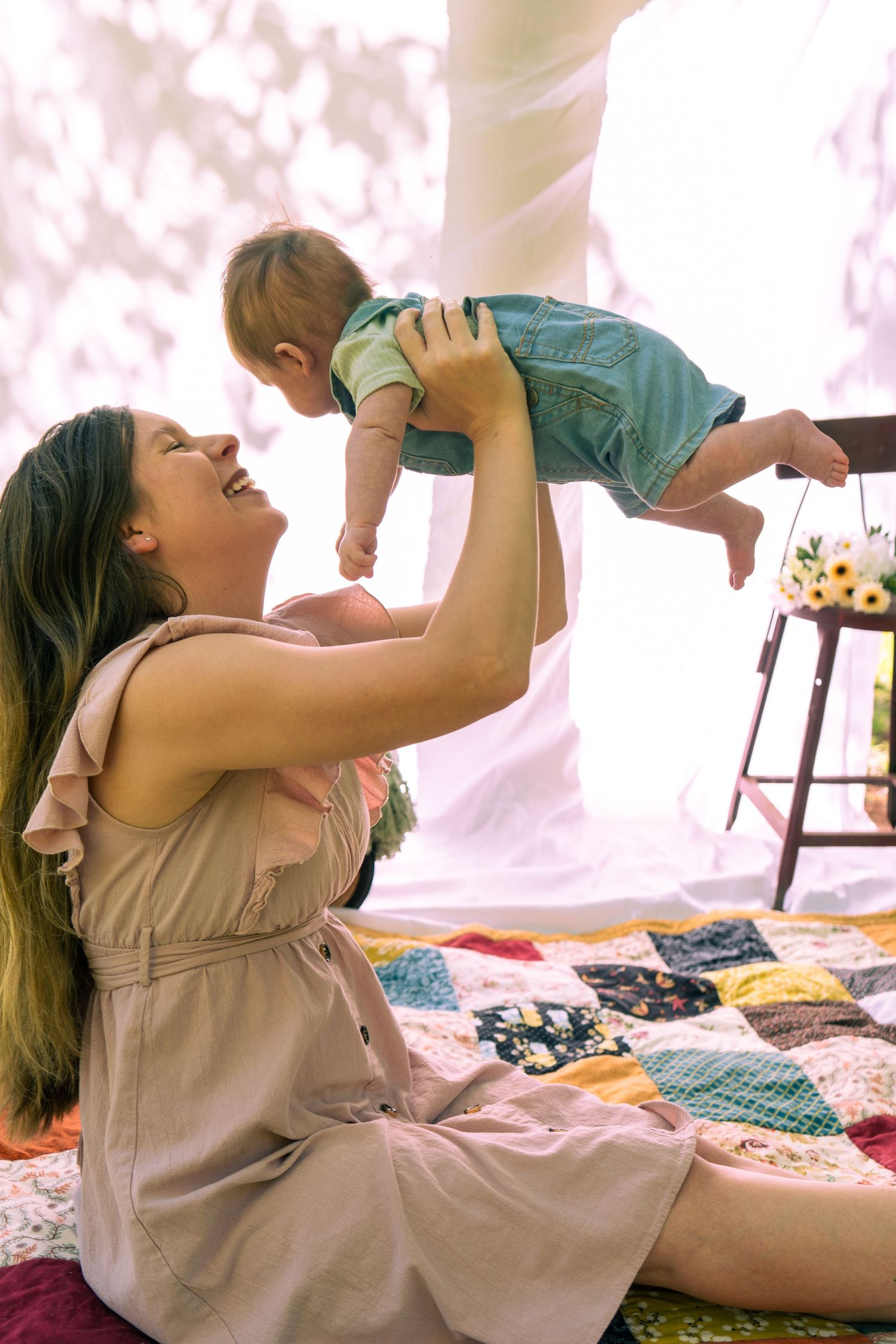 Joyful mother lifting and smiling at her baby on a colorful quilt indoors.