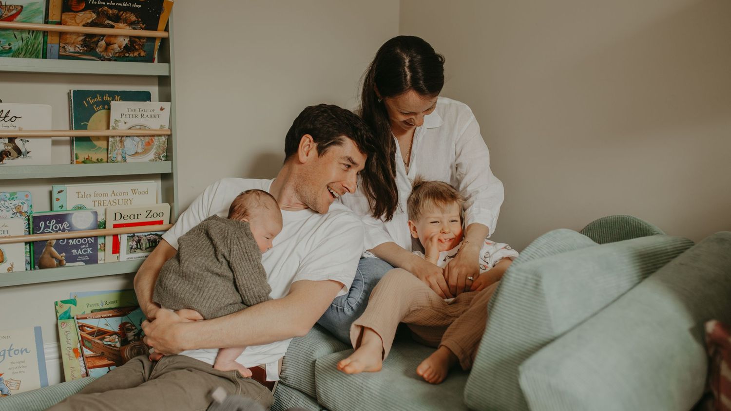 Happy family with newborn baby and toddler sitting on couch near bookshelf in cozy nursery room.