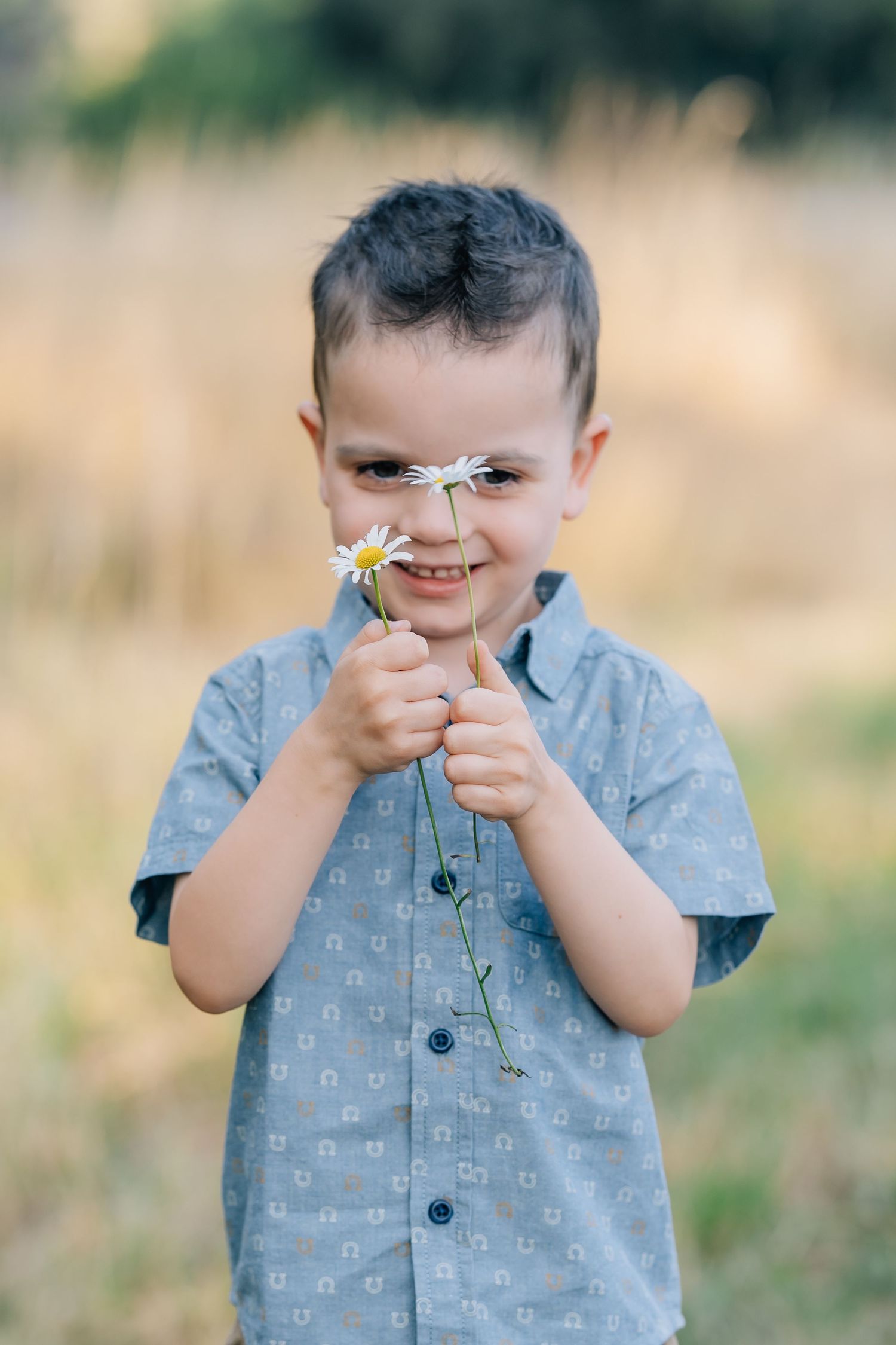 A smiling young boy in a blue shirt holds white daisy flowers up to his face outdoors in a sunny field.