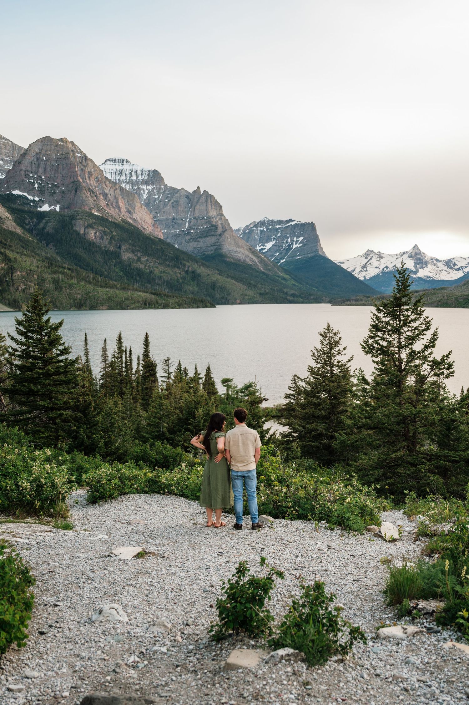Couple stands together overlooking Saint Mary Lake with dramatic Rocky Mountains and evergreen trees at Glacier National Park.