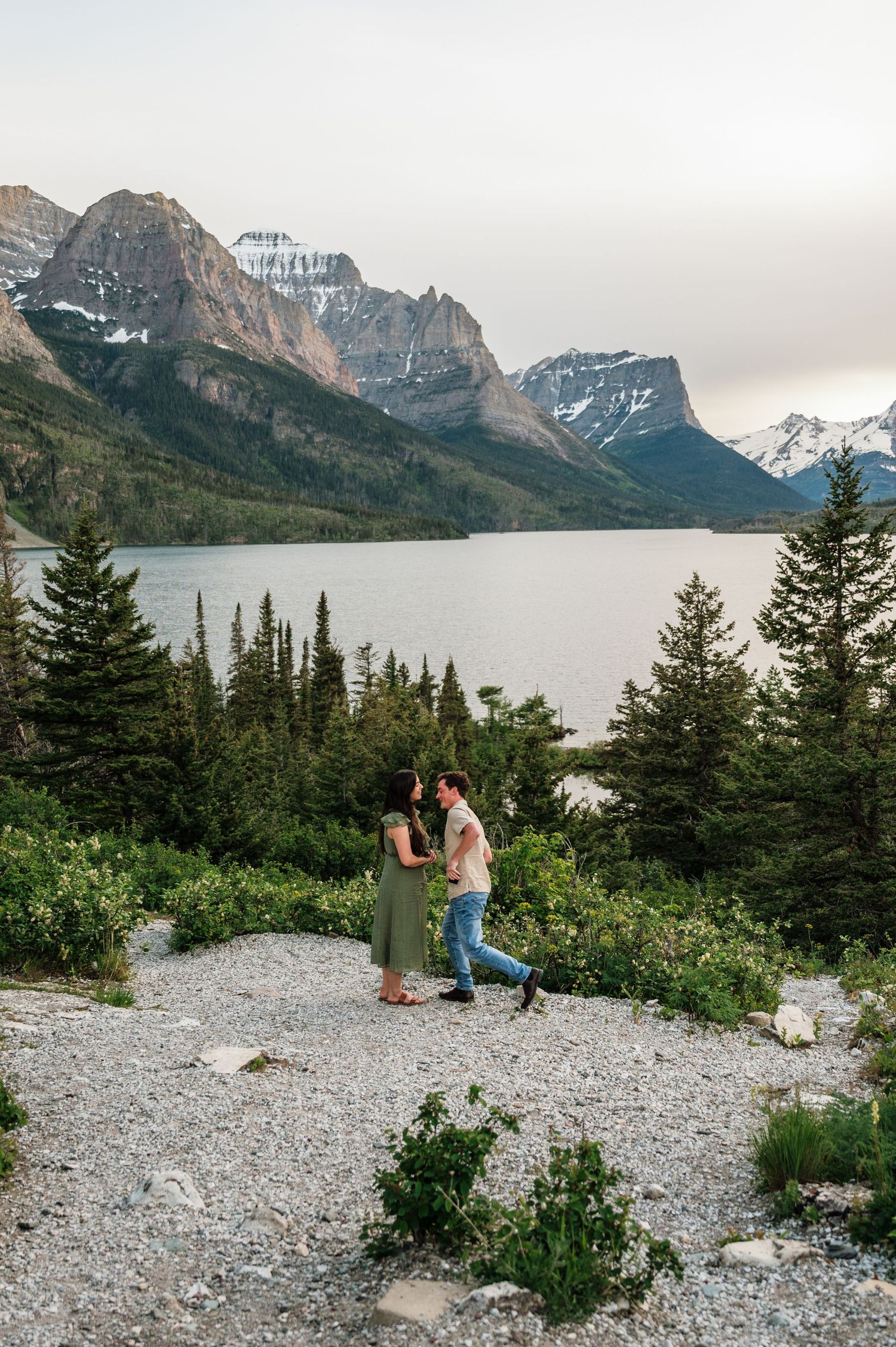 Couple holds hands and gazes at each other near Saint Mary Lake with snow-capped mountains at Glacier National Park.