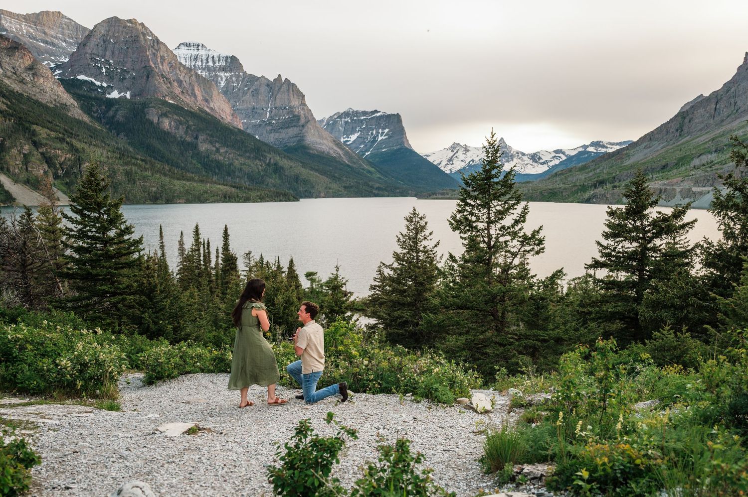 Couple sharing a romantic proposal moment on a rocky overlook with stunning mountain lake views at Glacier National Park.