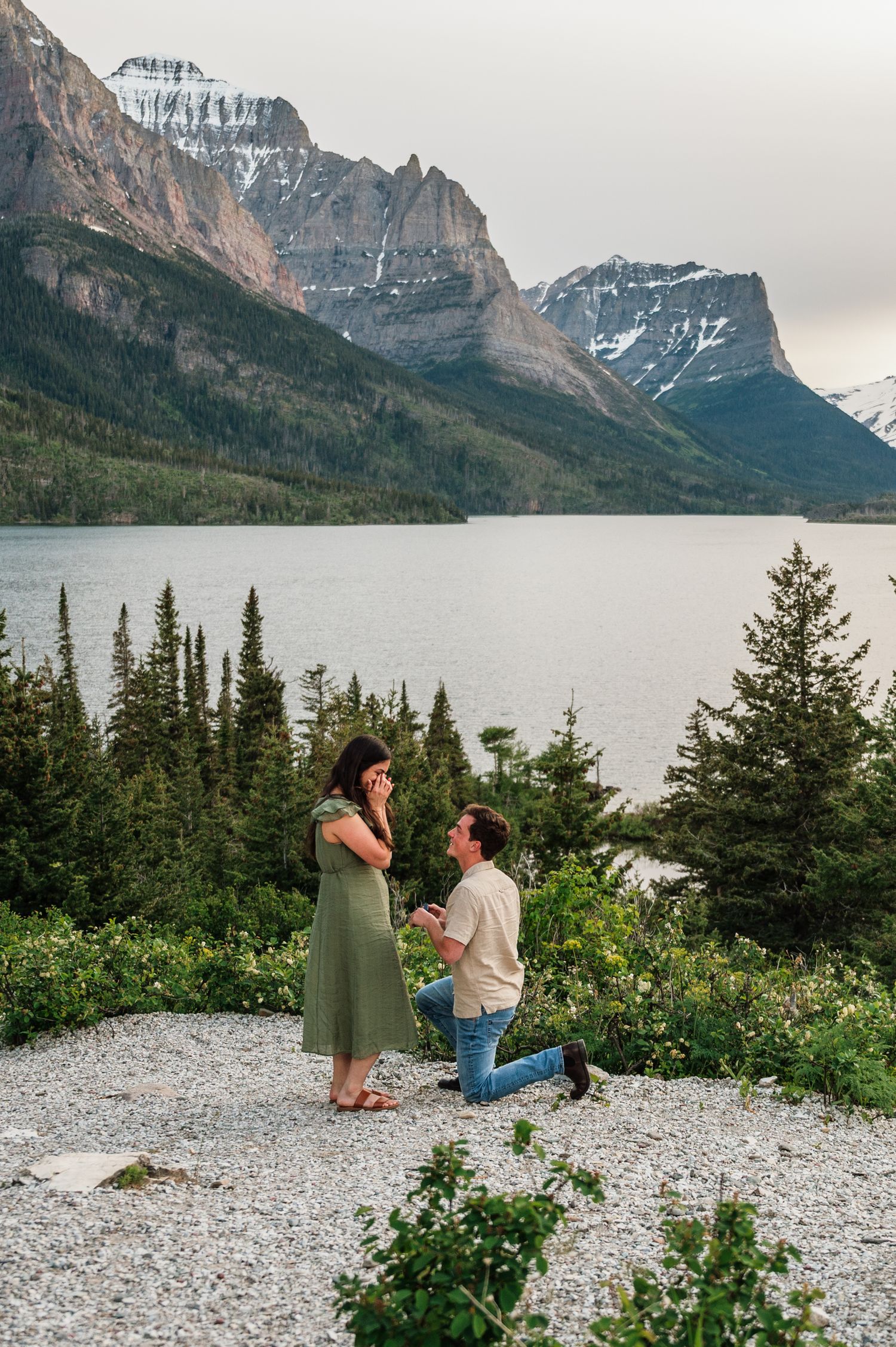 Man proposes to woman on one knee by a mountain lake with snow-capped peaks and evergreen trees in background.