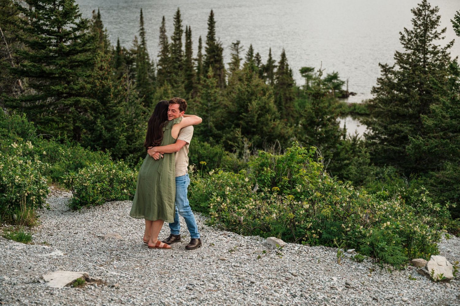 Couple embracing on a rocky path surrounded by lush greenery and pine trees near a lake.