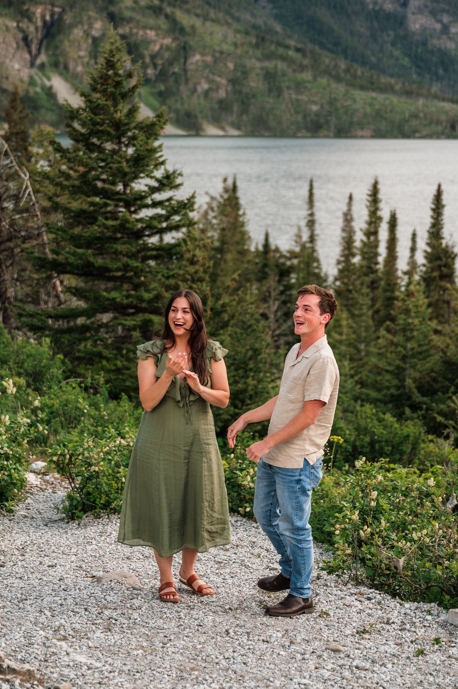 A couple laughing together on a scenic mountain trail with a lake and evergreen trees in the background.