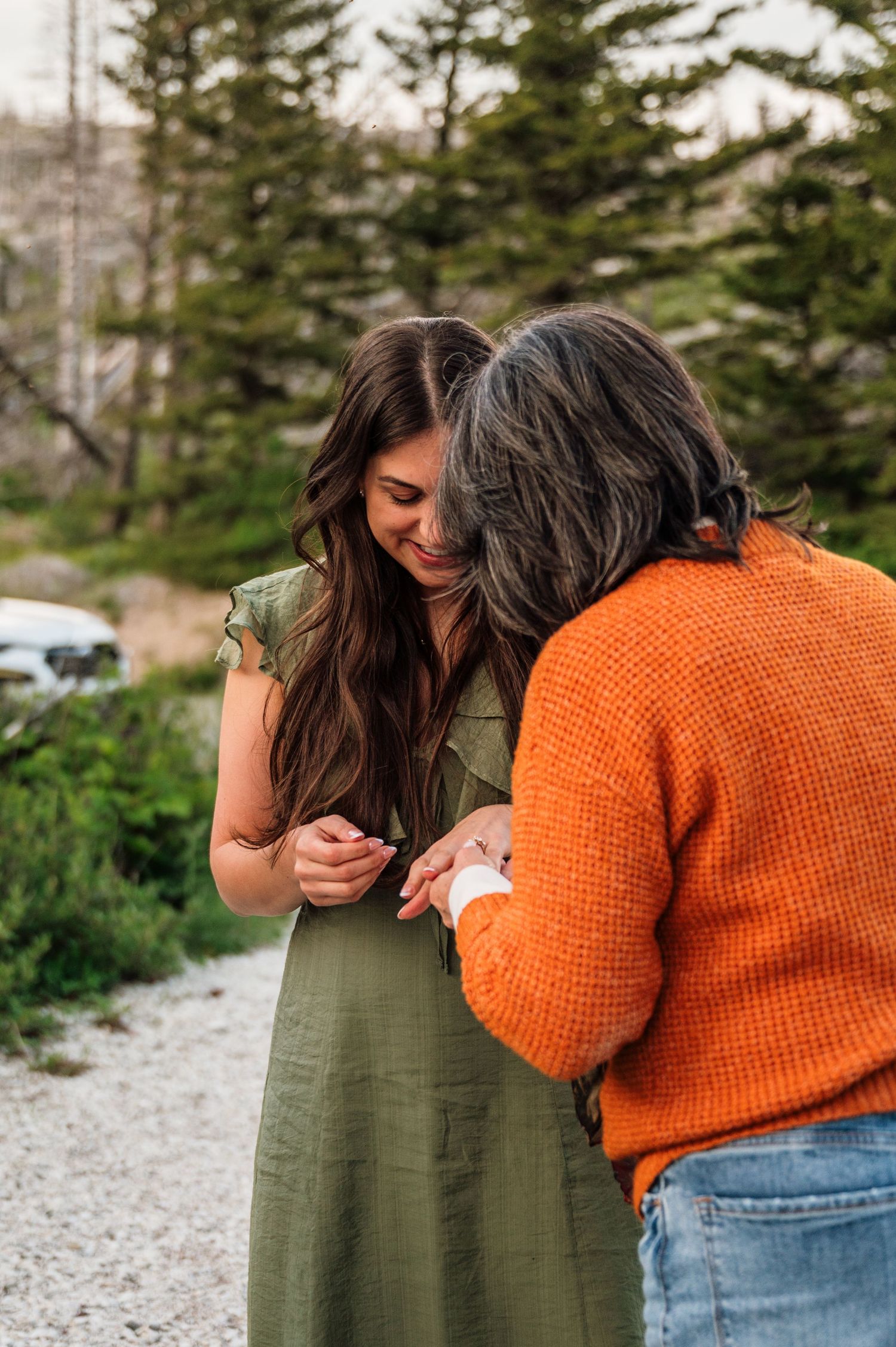 Two women share a joyful moment outdoors, one in green dress and other in orange sweater, possibly viewing an engagement ring.