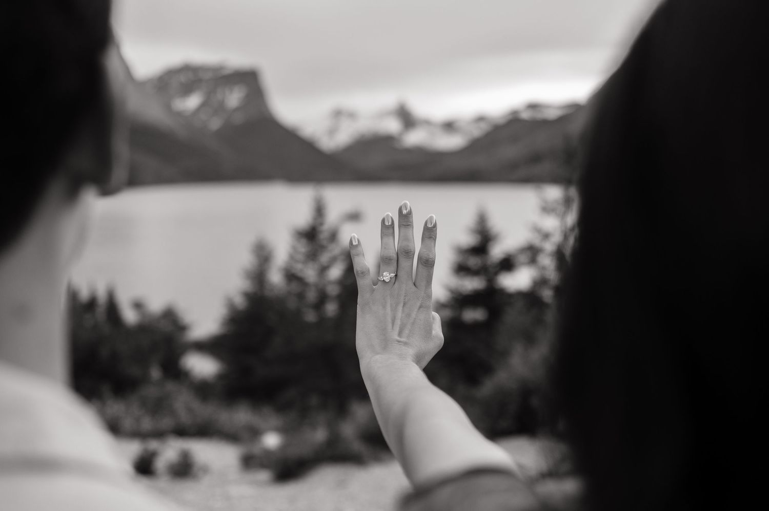 Black and white photo of a hand with engagement ring raised against mountain lake landscape.