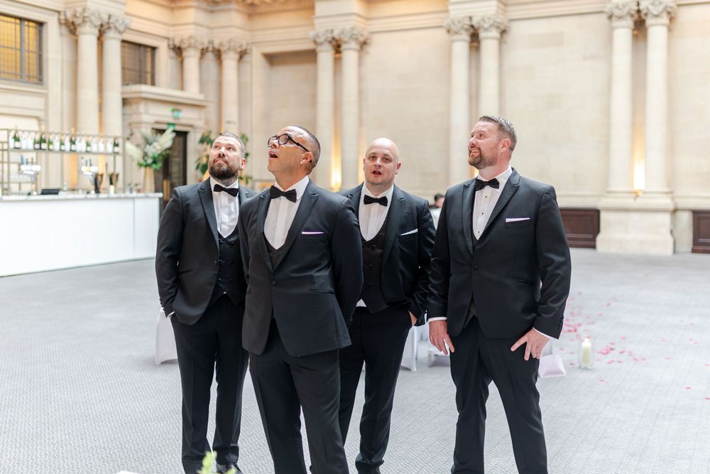 Four groomsmen in black tuxedos with bow ties stand together outdoors at an elegant wedding venue.