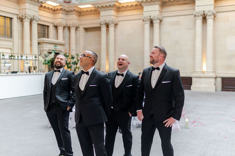 Four groomsmen in black tuxedos with bow ties laughing together in an elegant wedding venue.