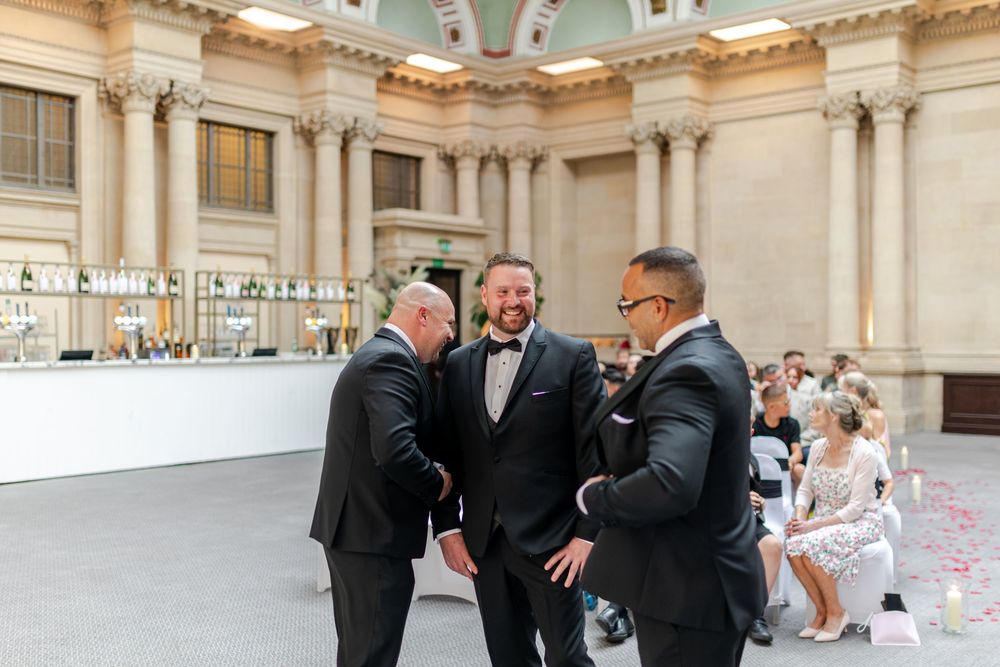 Three men in tuxedos share a joyful moment at an elegant wedding venue with grand architecture.