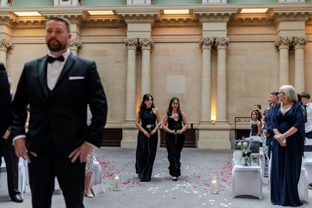Bridesmaids in black dresses walk down an aisle at an elegant outdoor wedding ceremony with floral decor.