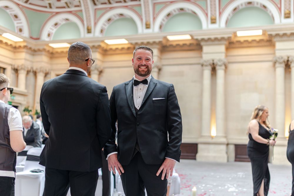 Two grooms in black tuxedos share a joyful moment at their elegant wedding venue with ornate architecture.