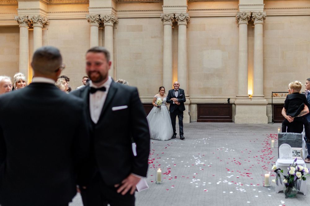 Groom watches bride walk down the aisle at an elegant outdoor wedding ceremony with rose petals.