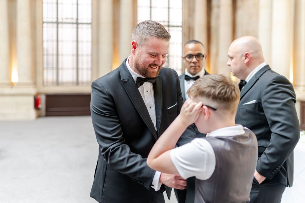 Groom in black tuxedo shares emotional moment with young boy at wedding ceremony, guests watching in background.