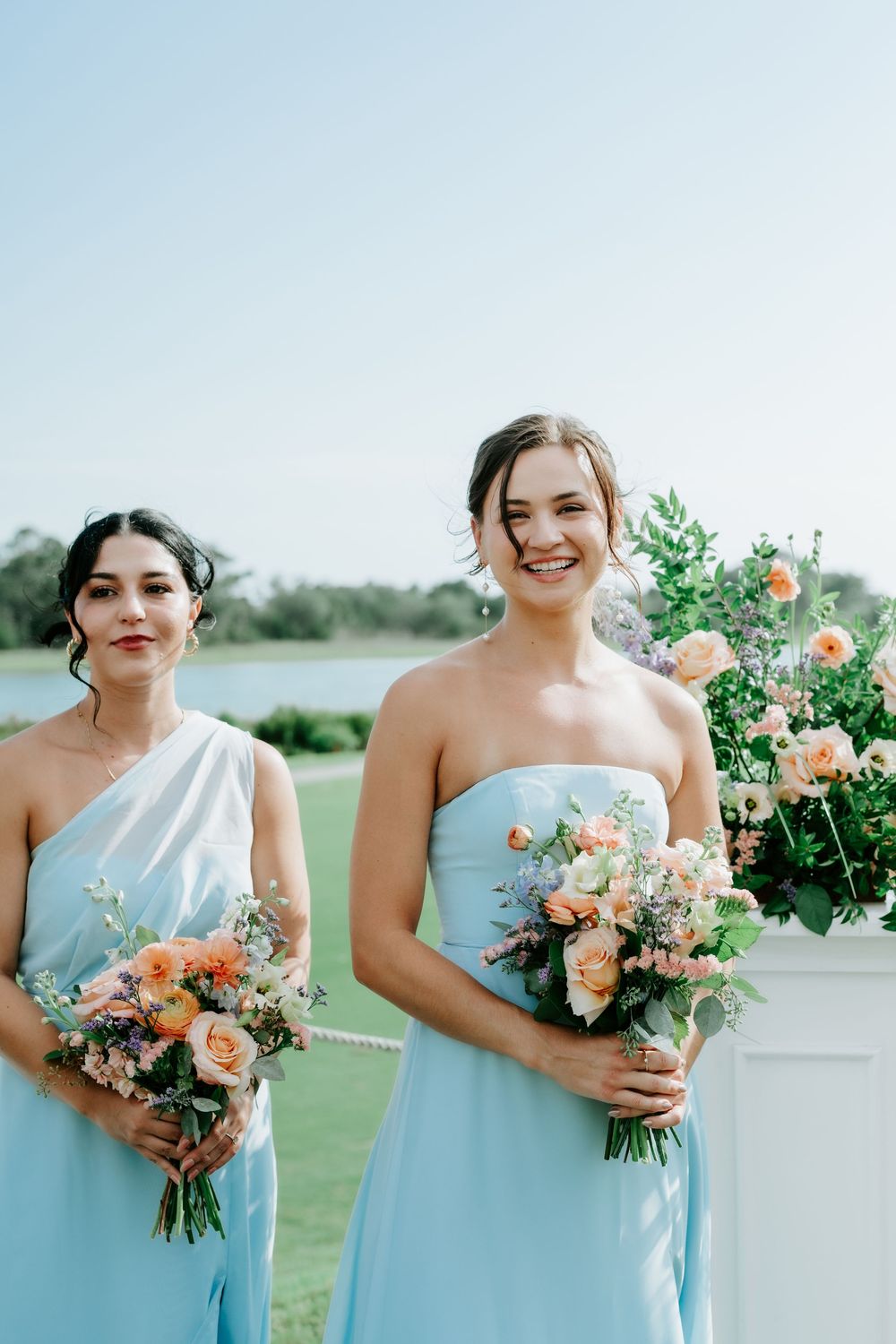 Two bridesmaids in light blue dresses holding peach floral bouquets smile outdoors at a wedding ceremony.