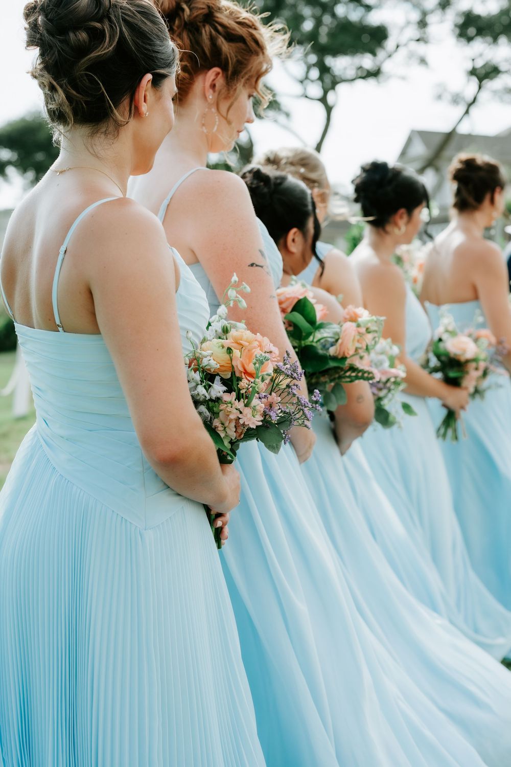 Bridesmaids in light blue dresses holding floral bouquets stand in a row outdoors at a wedding ceremony.