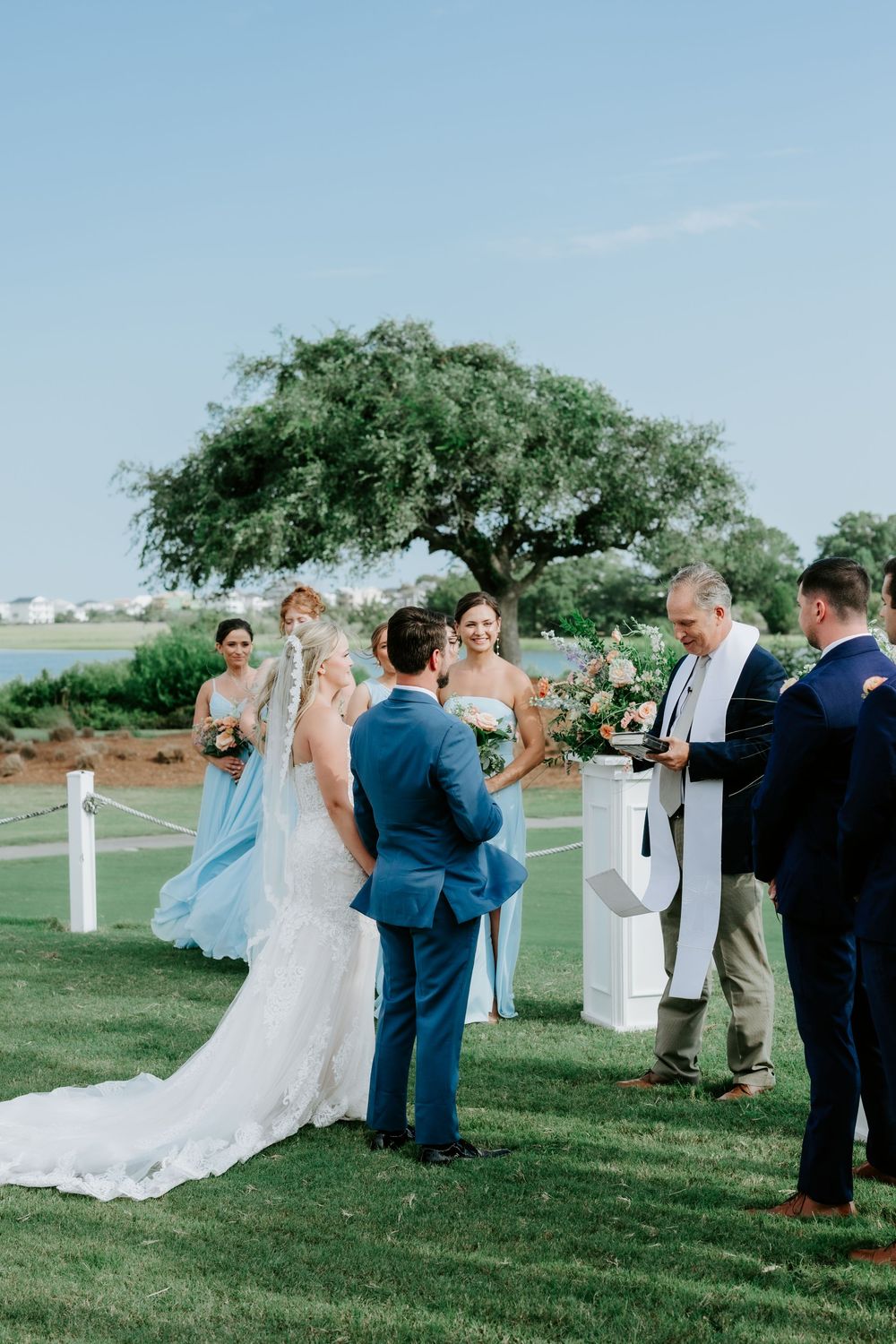 Outdoor wedding ceremony on green lawn with bride in lace gown, groom in blue suit, bridesmaids in light blue dresses.