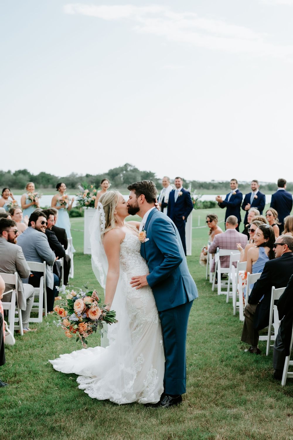 Bride and groom share first kiss at outdoor wedding ceremony on green lawn with guests seated in white chairs.