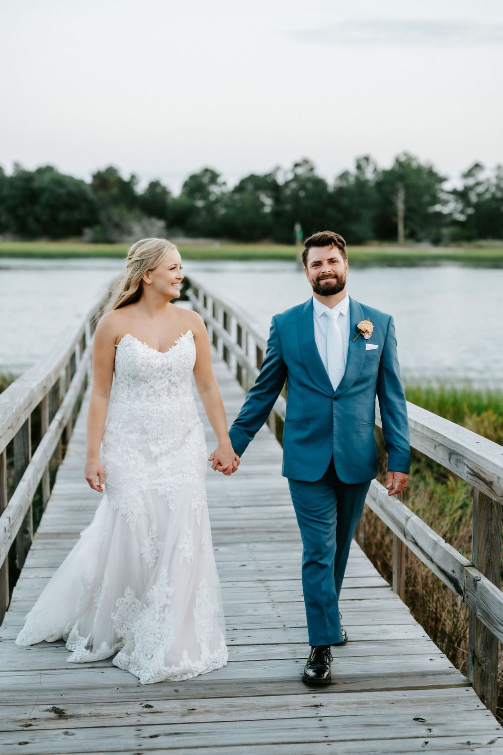 Bride and groom hold hands walking on a wooden dock by a serene waterway, she in lace gown, he in blue suit.