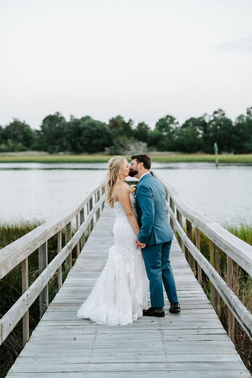 Newlyweds share a romantic kiss on a wooden dock overlooking a calm marsh, bride in lace gown and groom in blue suit.
