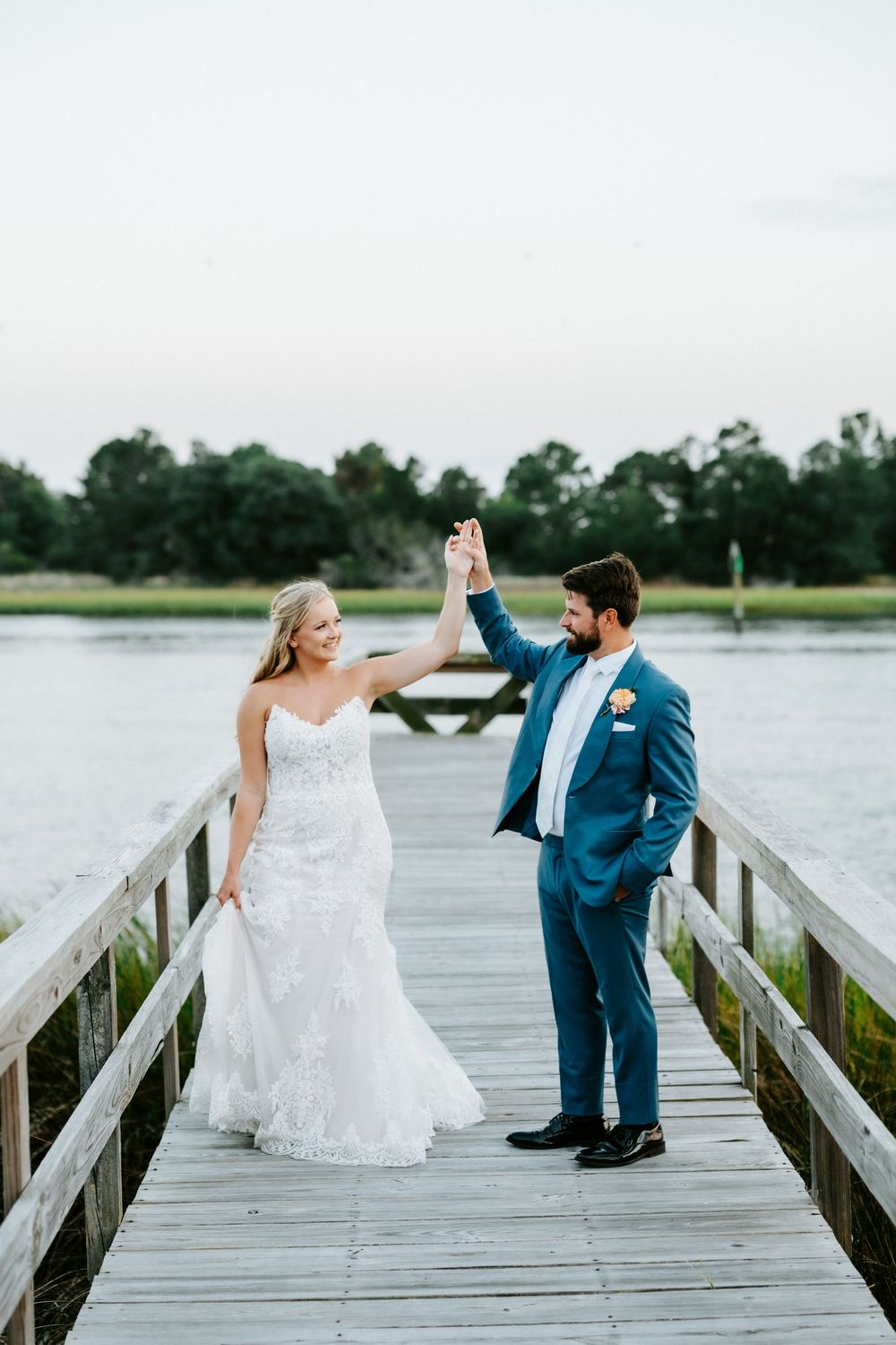 Bride and groom holding hands on a wooden dock by the water, groom in blue suit, bride in lace mermaid gown.